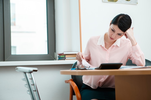 Young businesswoman sitting at workplace and reading paper in office