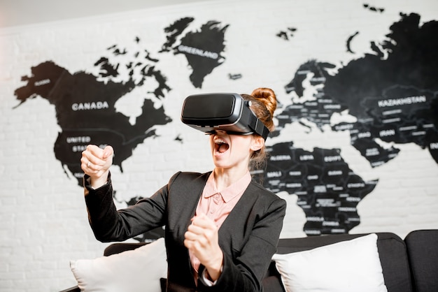 Young businesswoman trying virtual reality glasses sitting indoors with world map on the background