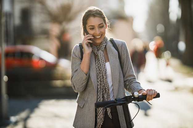 A young businesswoman using a smartphone while traveling with an electric scooter through the city.