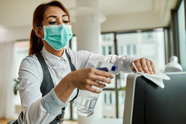 Young businesswoman wearing face mask and cleaning her computer while working in the office during virus epidemic