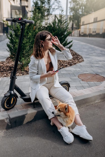 Young businesswoman in white suit sitting on electric scooter and using phone during walking with Welsh Corgi Pembroke dog in city