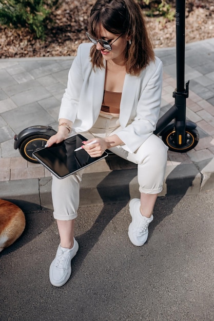 Young businesswoman in white suit sitting on electric scooter and working on digital tablet during walking with Welsh Corgi Pembroke dog in city