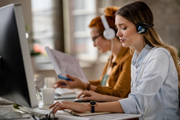 Young businesswoman with headset working on the computer in the office Her colleague is in the background