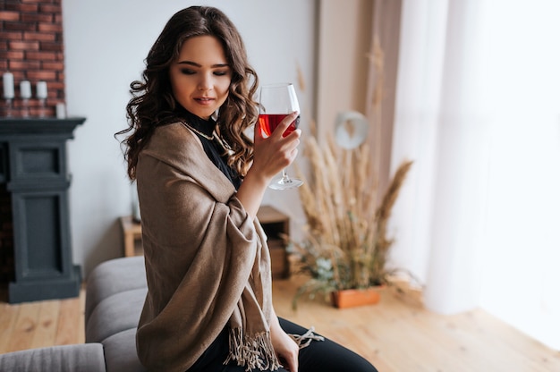 Young businesswoman work at home. Hold glass of red wine in hand. Beautiful model in evening dress stand at window. Alone in room.