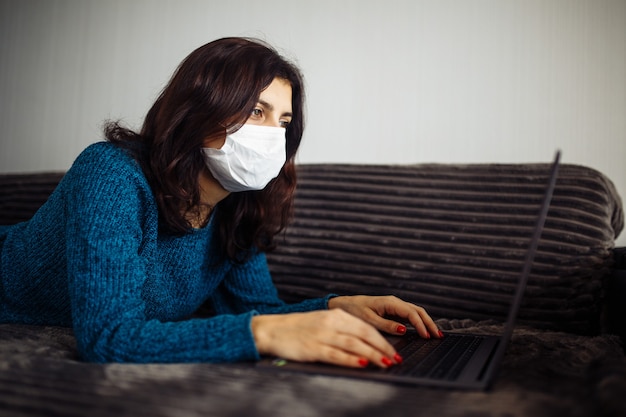 Young businesswoman working from home during quarantine due to coronavirus pandemia. Beautiful girl stays home wearing medical mask and typing on a laptop. Covid-19 epidemia worldwide concept.