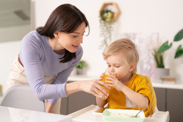 Young careful mother holding glass of orange juice while bending by her cute little son drinking it by small dinner table during breakfast