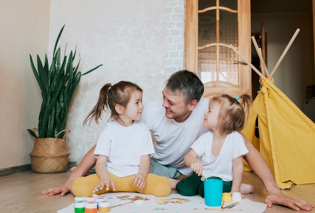 A young caring father learns to paint with his young daughters, a man lies on the floor and teaches girls to draw flowers, home furnishings.