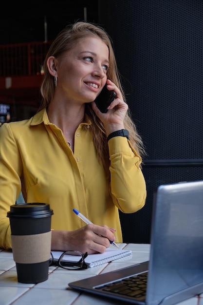 Young cauasian pretty woman sitting with laptop at cafe calling by smartphone