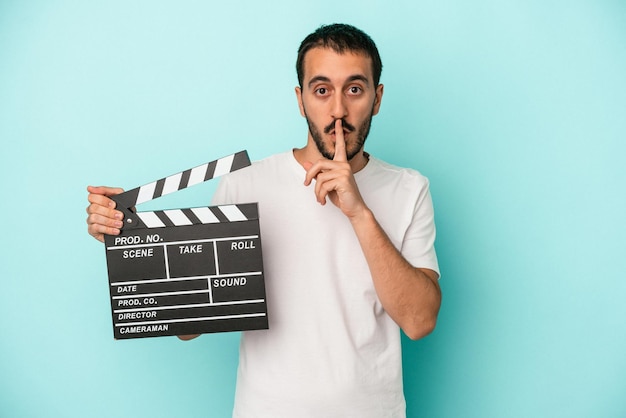 Young caucasian actor man holding clapperboard isolated on blue background keeping a secret or asking for silence.