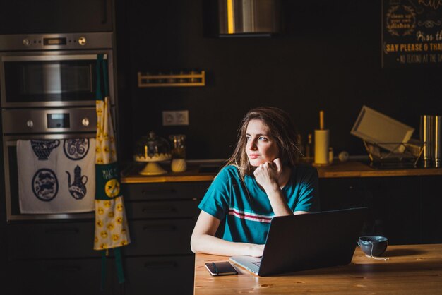 Young caucasian adult sitting in her kitchen with her laptop working from home looking away.