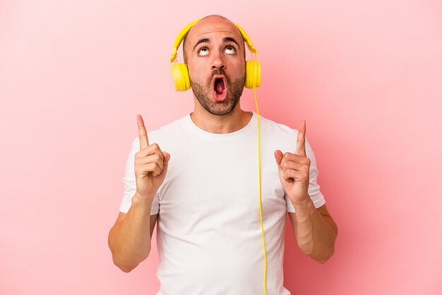 Young caucasian bald man listening to music isolated on pink background  pointing upside with opened mouth.