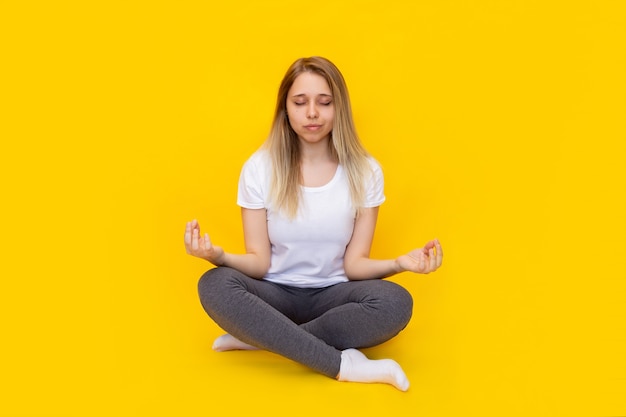 Photo a young caucasian beautiful blonde woman in a white tshirt and grey leggings sitting in the lotus position meditating isolated on a bright color yellow wall