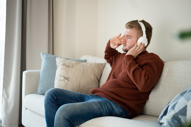 Young caucasian blond curly hair man listening to music with big white handsfree headphones sitting on sofa at home on neutral white background Handsome adult guy portrait lifestyleCopyspace