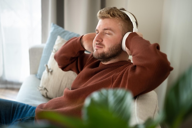 Young caucasian blond curly hair man listening to music with big white handsfree headphones sitting on sofa at home on neutral white background Handsome adult guy portrait lifestyleCopyspace