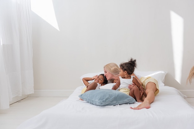 Young Caucasian blond mother plays, lies on white bed with African American daughter son