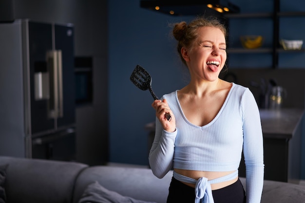 Young caucasian blonde woman holding a black spatula sticks out tongue with her eyes closed laughing