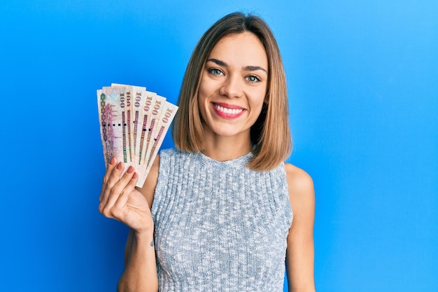 Young caucasian blonde woman holding saudi arabia riyal banknotes looking positive and happy standing and smiling with a confident smile showing teeth