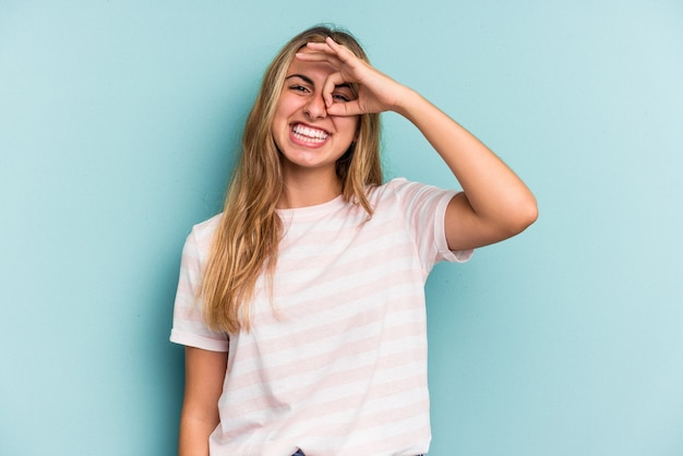 Young caucasian blonde woman isolated on blue background  excited keeping ok gesture on eye.