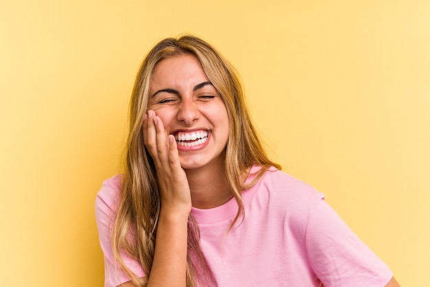 Young caucasian blonde woman isolated on yellow background  laughs happily and has fun keeping hands on stomach.