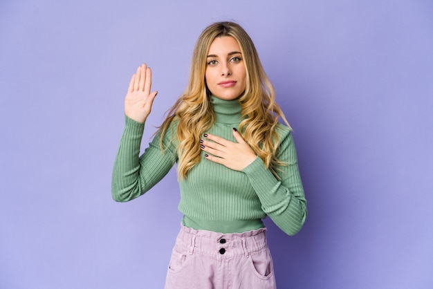 Young caucasian blonde woman taking an oath, putting hand on chest.