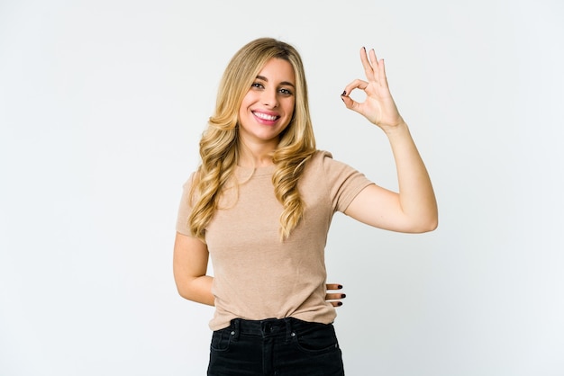 Young caucasian blonde woman winks an eye and holds an okay gesture with hand.