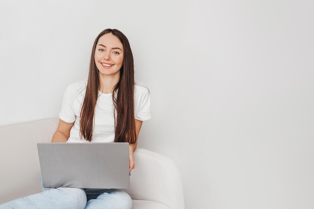 Young caucasian brunette woman smiling at camera and sitting with laptop on sofa