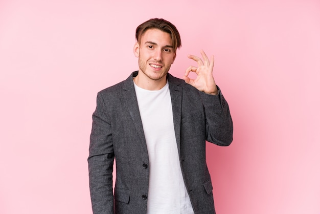 Young caucasian business man posing isolated winks an eye and holds an okay gesture with hand.