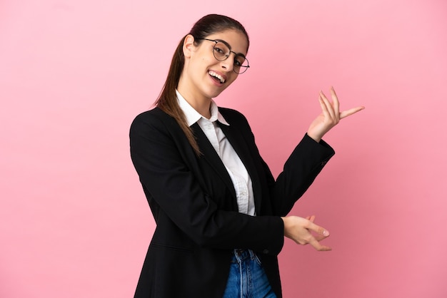 Young caucasian business woman isolated on pink background extending hands to the side for inviting to come