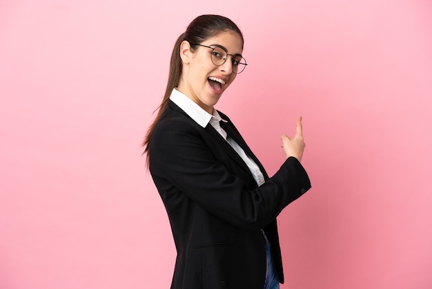 Young caucasian business woman isolated on pink wall pointing back