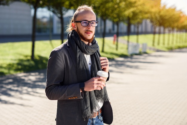 Young caucasian businessman with backpack and cup of coffee outdoors near modern business center.