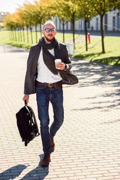 Young caucasian businessman with backpack and cup of coffee outdoors near modern business center.