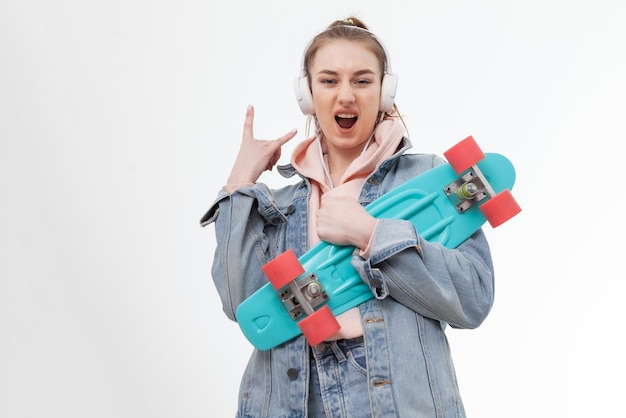 Young caucasian cheerful hipster woman in denim clothes with penny board and headphones listen music on white background