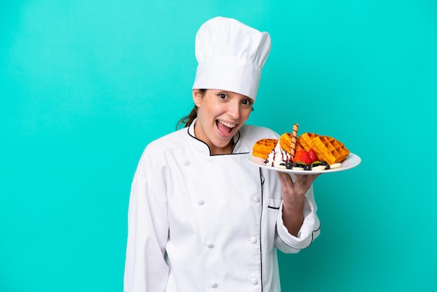 Young caucasian chef woman holding waffles isolated on blue background with surprise and shocked facial expression