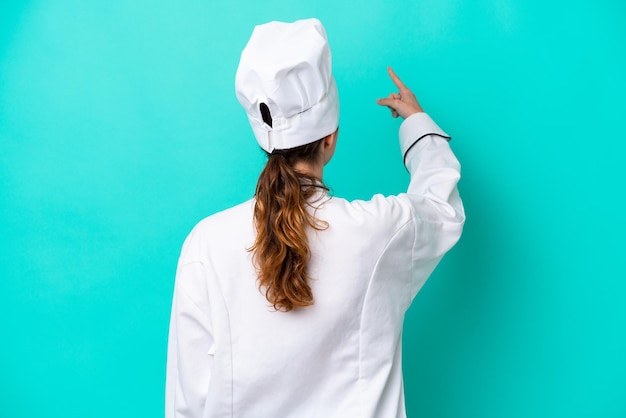 Young caucasian chef woman isolated on blue background pointing back with the index finger