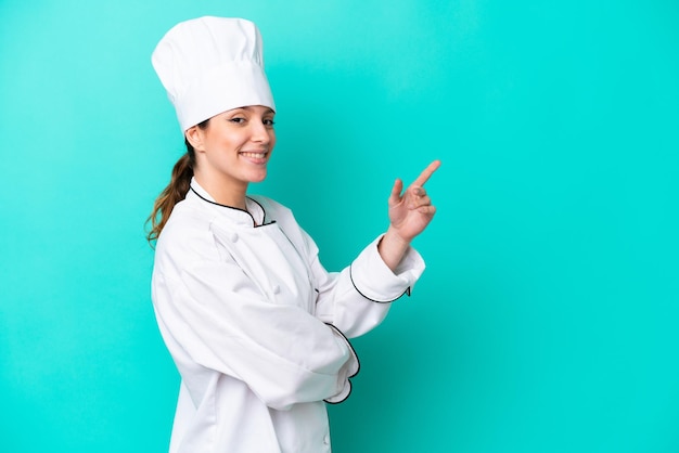 Young caucasian chef woman isolated on blue background pointing finger to the side