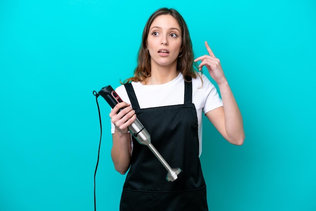 Young caucasian cooker woman using hand blender isolated on blue background thinking an idea pointing the finger up