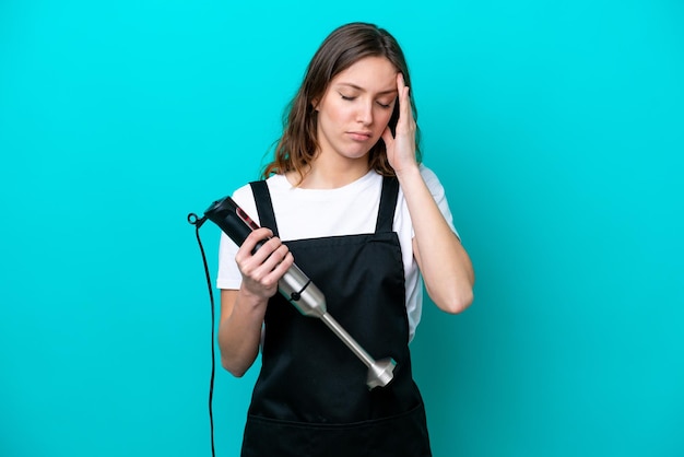 Young caucasian cooker woman using hand blender isolated on blue background with headache