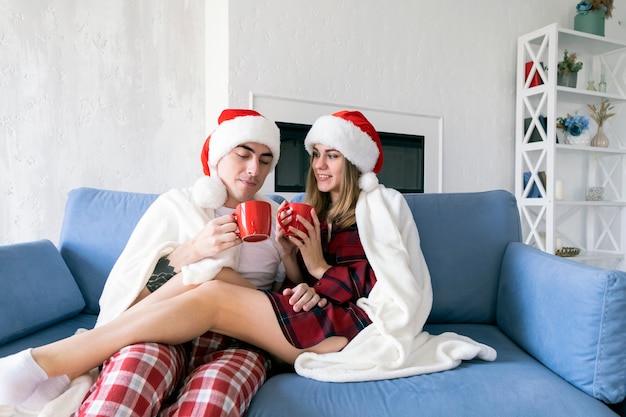 Young Caucasian couple drinking hot tea or cocoa from red cups while sitting at home on the sofa wearing santa hats and pajamas wrapped in a white blanket