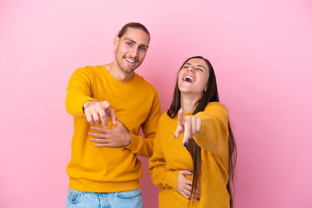 Young caucasian couple isolated on pink background pointing with finger at someone and laughing a lot