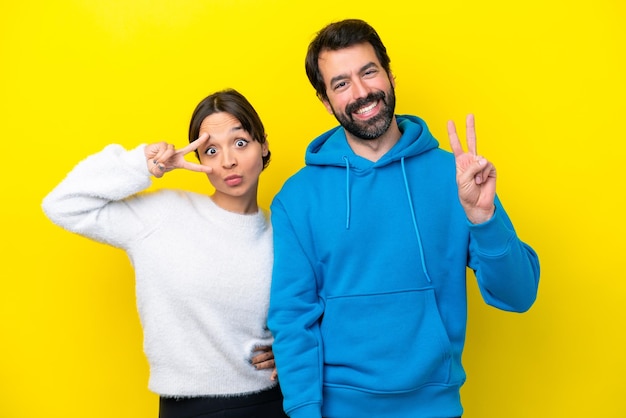 Young caucasian couple isolated on yellow background smiling and showing victory sign