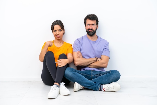 Young caucasian couple sitting on the floor isolated on white background annoyed angry in furious gesture