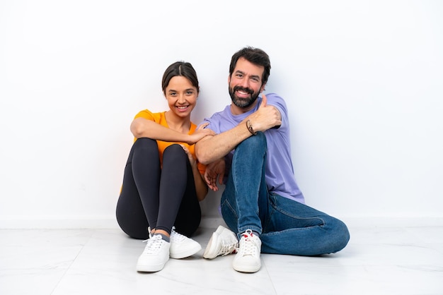 Young caucasian couple sitting on the floor isolated on white background giving a thumbs up gesture