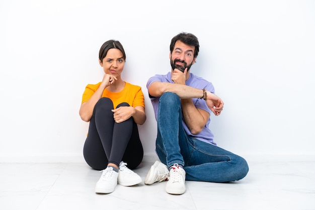 Young caucasian couple sitting on the floor isolated on white background having doubts and with confuse face expression