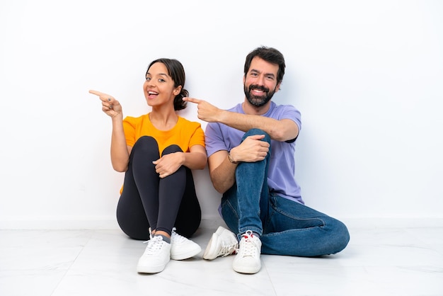 Young caucasian couple sitting on the floor isolated on white background pointing finger to the side and presenting a product