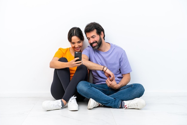 Young caucasian couple sitting on the floor isolated on white background sending a message or email with the mobile