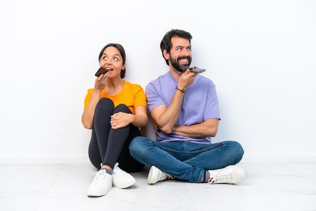 Young caucasian couple sitting on the floor isolated on white background sending a message voice or email with the mobile