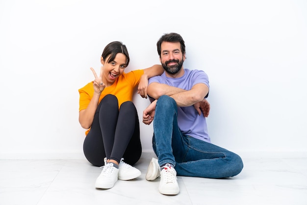 Young caucasian couple sitting on the floor isolated on white background smiling and showing victory sign