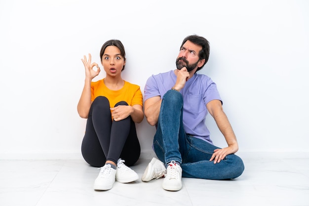 Young caucasian couple sitting on the floor isolated on white background standing and thinking an idea