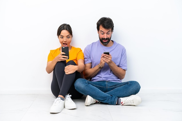 Young caucasian couple sitting on the floor isolated on white background Surprised and sending a message or email with the mobile