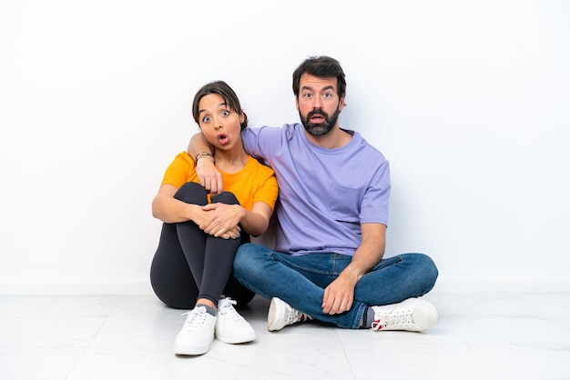 Young caucasian couple sitting on the floor isolated on white background with surprise facial expression